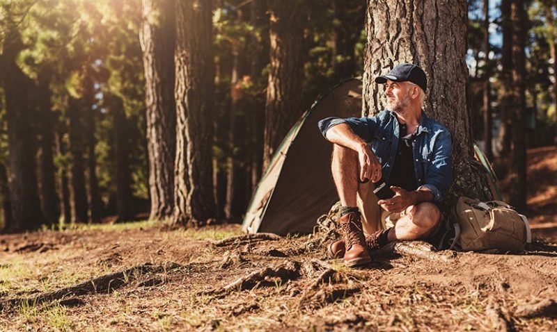 Portrait of senior man sitting by a tree with a tent in background. Mature man sitting at a campsite.